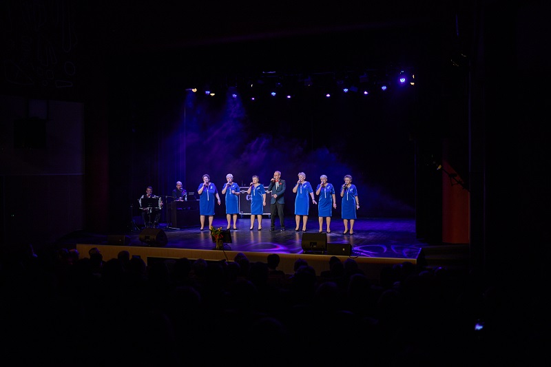Na scenie stoi sześć kobiet i mężczyzna z mikrofonem w ręku, przy organach i z akordeonem siedzi dwóch mężczyzn