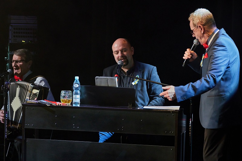 Na scenie trzech mężczyzn śpiewających do mikrofonów. Jeden stoi, dwóch siedzi przy akordeonie i organach