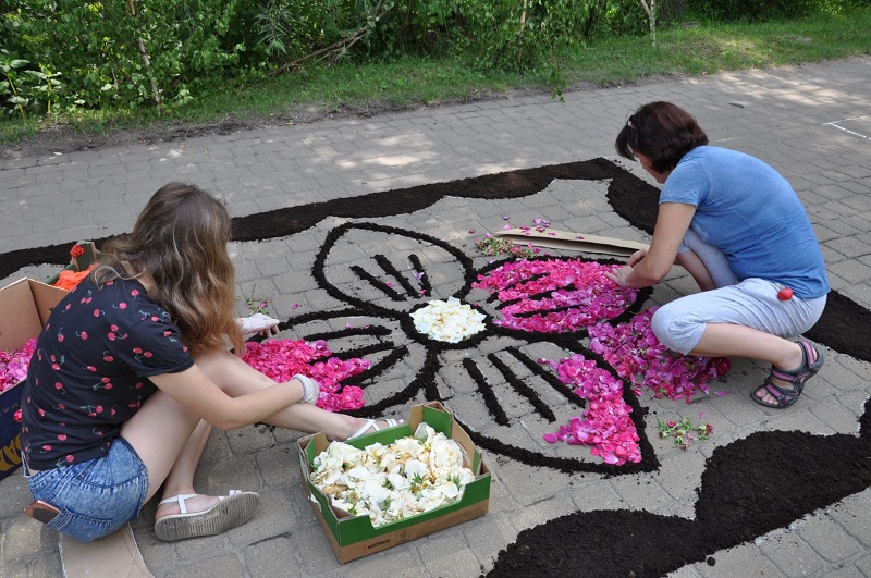  Dwie dziewczyny układają płatki kwiatów w środku dużego kwiata obramowanego czarnoziemem. W pudełkach stoją przygotowane do dekoracji pąki białych kwiatów.