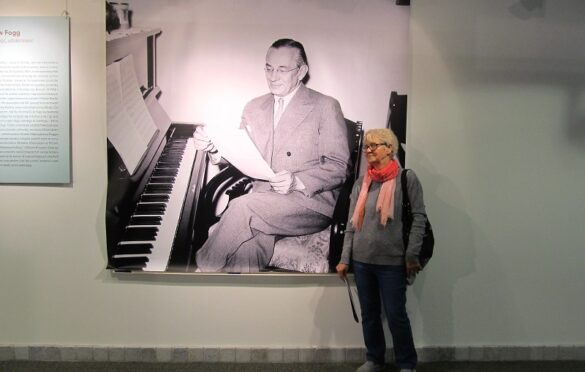 Przed dużym zdjęciem Mieczysława Fogga stoi kobieta. Na zdjęciu Fogg siedzi przy pianinie i trzyma w rękach kartkę.
