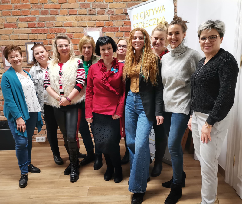 7.Grupa dziesięciu uśmiechniętych kobiet w sali z ceglaną ścianą w tle i banerem z napisem Inicjatywa Społeczna.