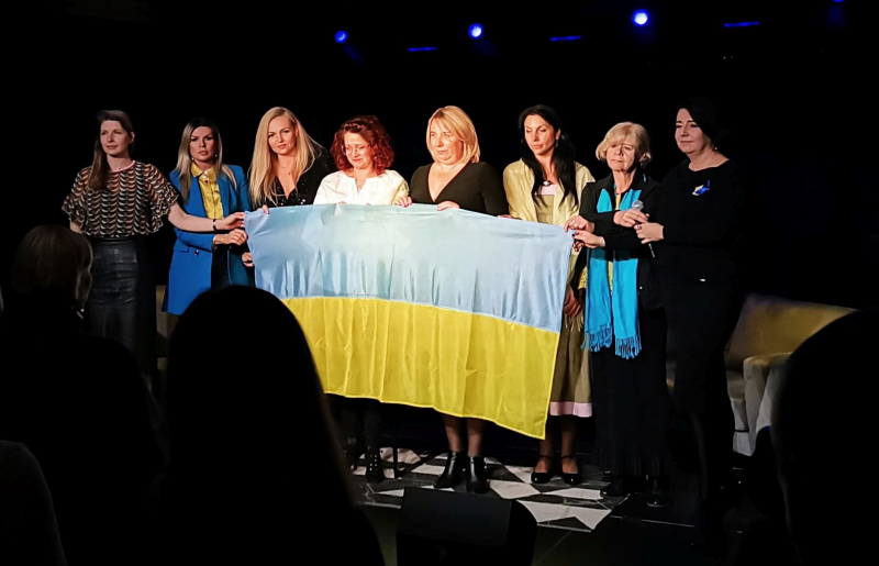 6. Grupa ośmiu kobiet trzyma przed sobą na scenie flagę Ukrainy.
