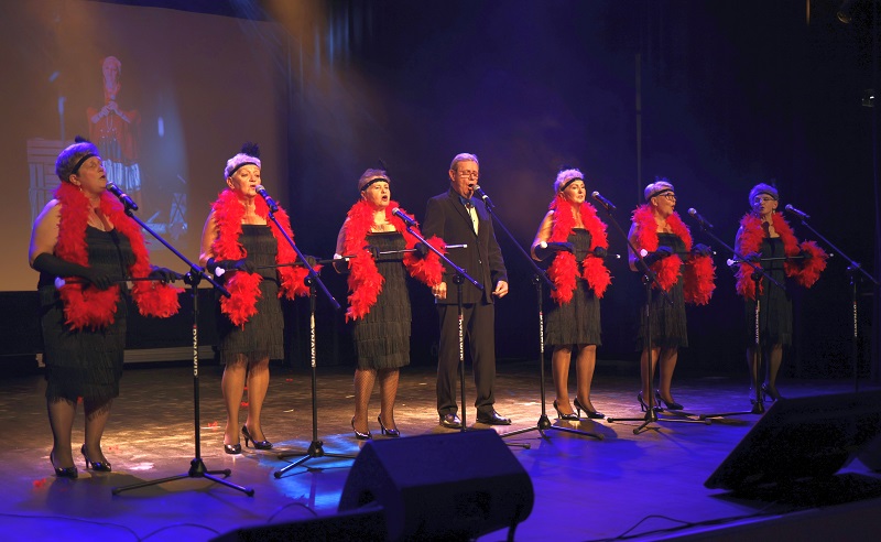 Na scenie grupa śpiewających osób-sześć pań w czarnych sukniach, z czerwonymi szalami, w środku mężczyzna w garniturze.