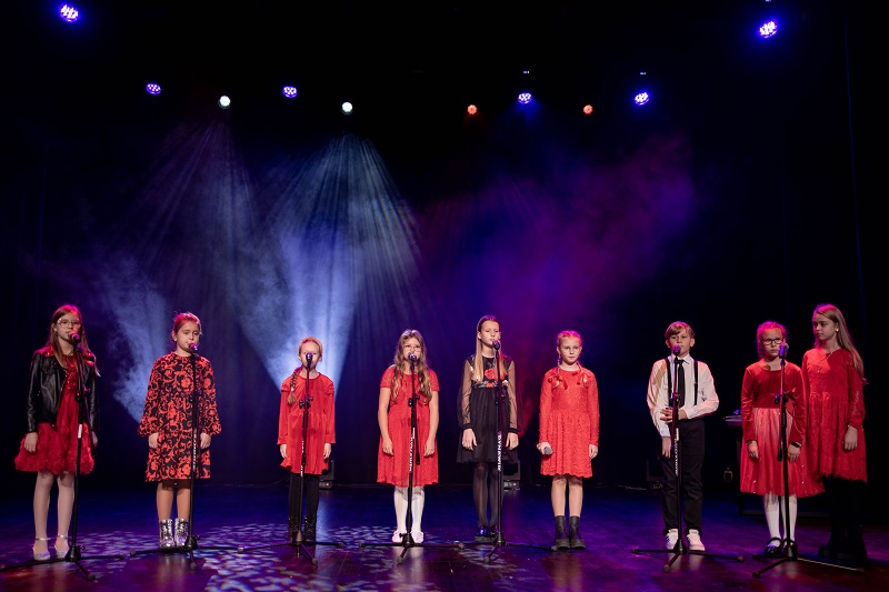 Na scenie przed mikrofonami stoi śpiewajaca ósemka dziewczynek i chłopak.