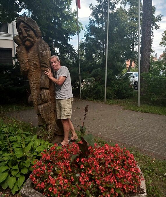 Mężczyzna przytula się do drewnianej rzeźby rybaka. Wokół różnokolorowe kwiaty i drzewa.
