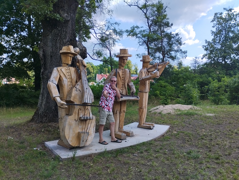Obok drewnianych rzeźb trzech muzyków stoi mężczyzna z wyciągniętą ręką i uniesionym kciukiem.