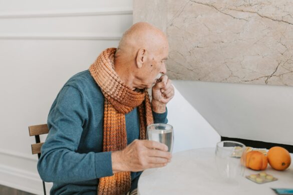 starszy mężczyzna siedzi przy stole, szklanką wody popija tabletkę.